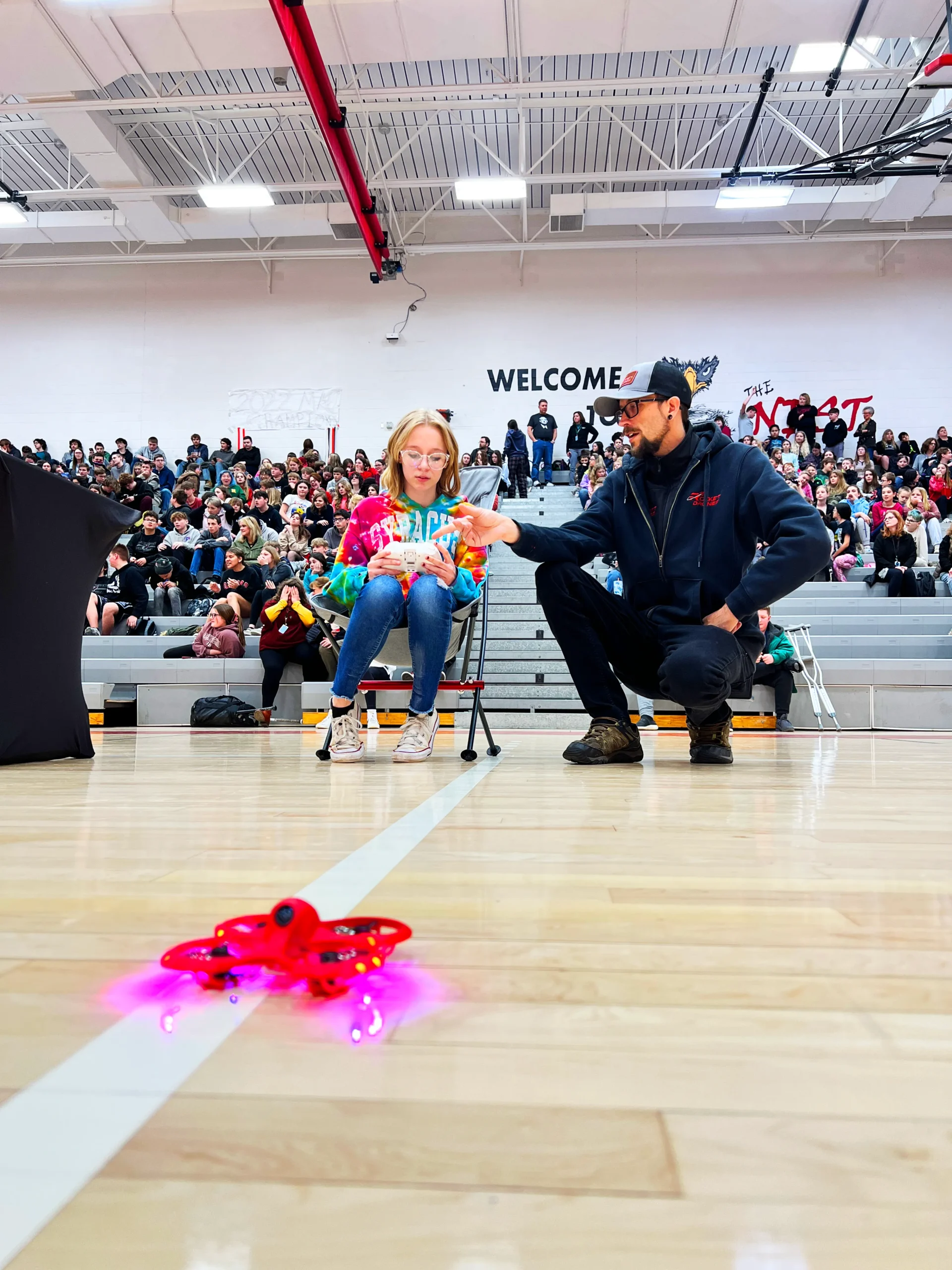A student learning to fly a Rocket Drones training drone in a school gym during a STEM education event, guided by an instructor.