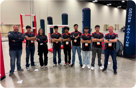 Drone racing students and instructors pose in front of Rocket Drones training gates inside an indoor competition arena.